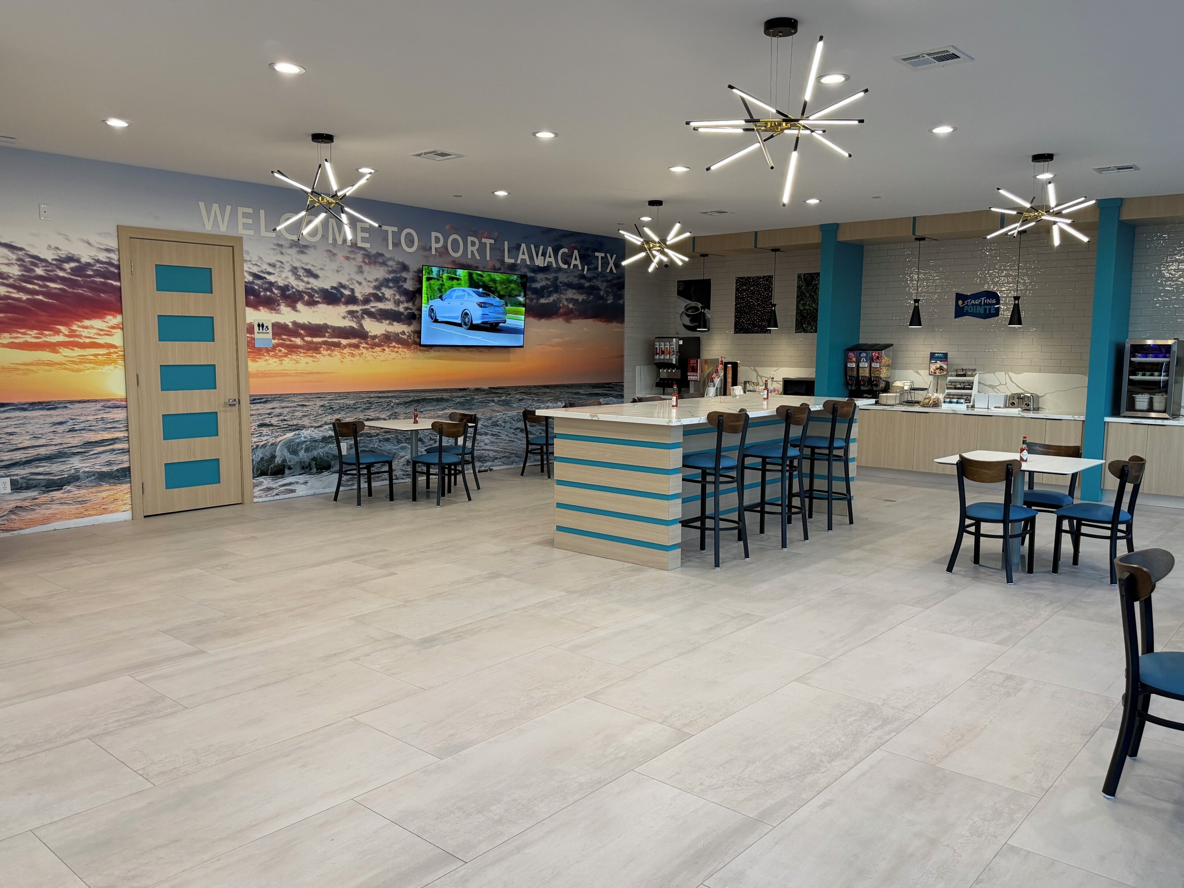 Un interior moderno con una zona de bar, asientos, un televisor mostrando un automóvil y un mural de un atardecer sobre el océano.