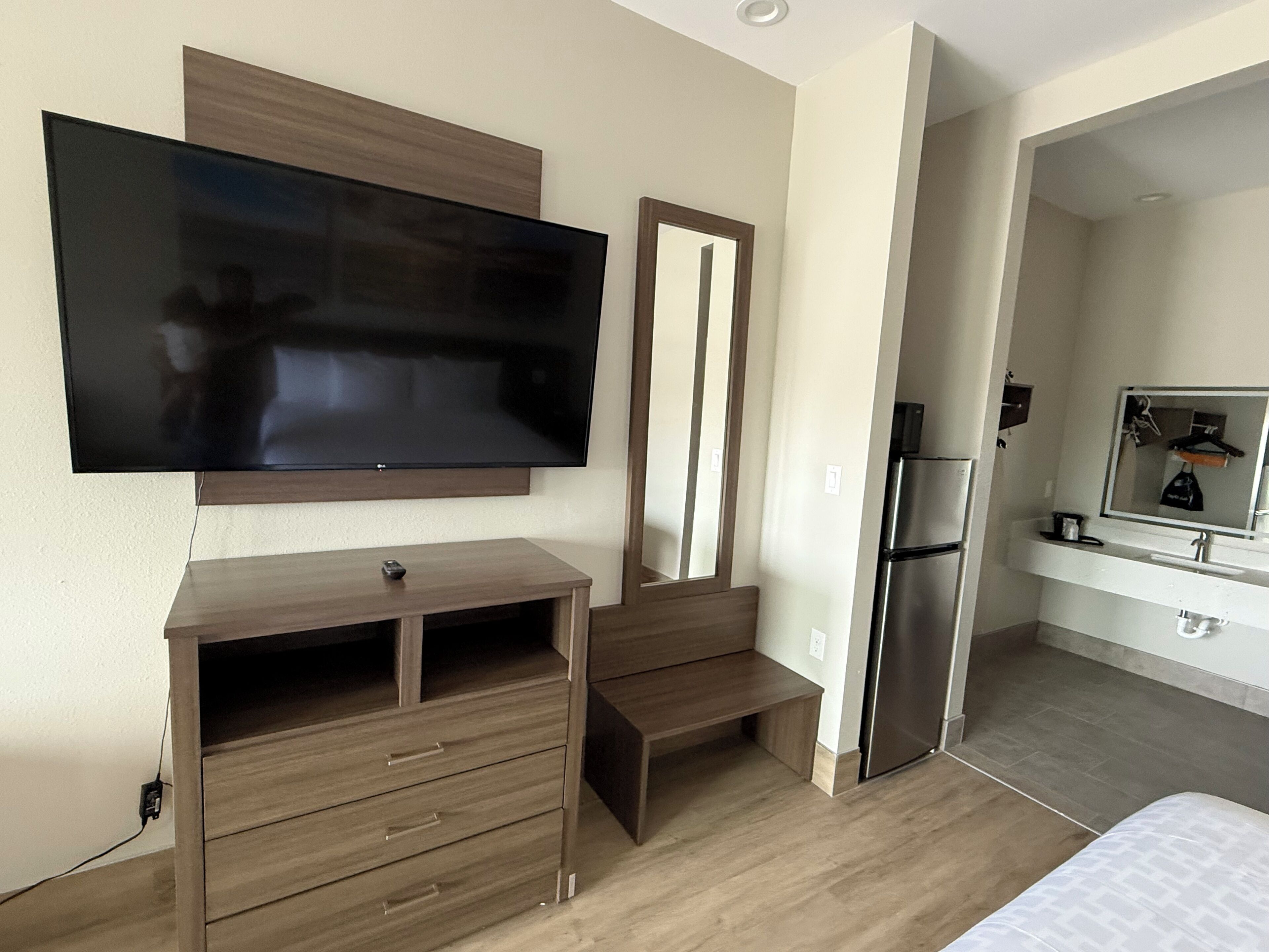 A modern hotel room with a flat-screen TV mounted on the wall, a wooden dresser with drawers, a built-in bench, and a refrigerator in the adjacent kitchenette.