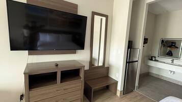 A modern hotel room with a flat-screen TV mounted on the wall, a wooden dresser with drawers, a built-in bench, and a refrigerator in the adjacent kitchenette.