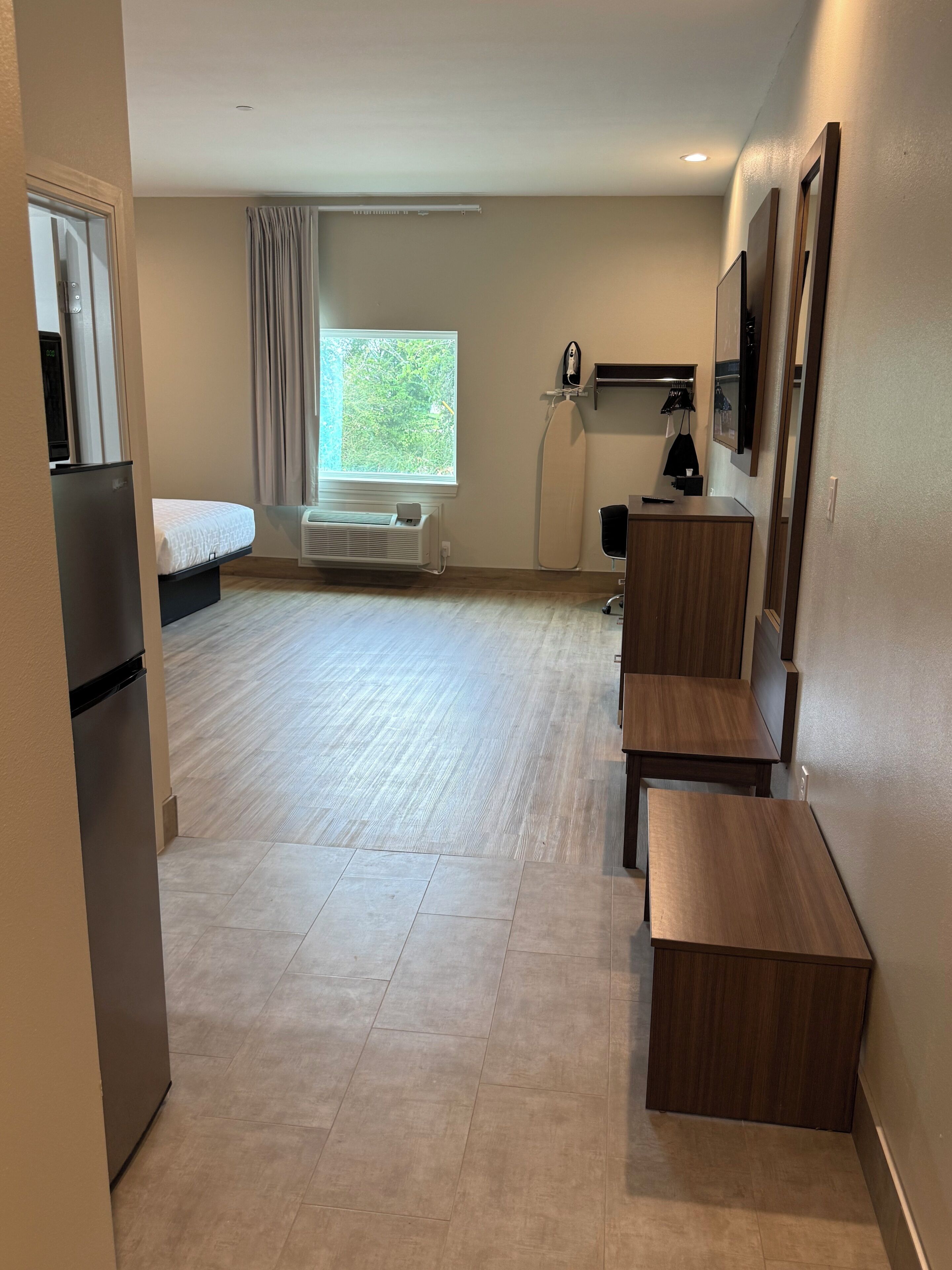 Une chambre d’hôtel de taille réduite comprenant un lit, un petit bureau, une chaise, un banc, une fenêtre avec des rideaux, un radiateur et une planche à repasser fixée au mur.