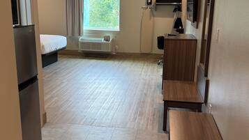 Une chambre d’hôtel de taille réduite comprenant un lit, un petit bureau, une chaise, un banc, une fenêtre avec des rideaux, un radiateur et une planche à repasser fixée au mur.