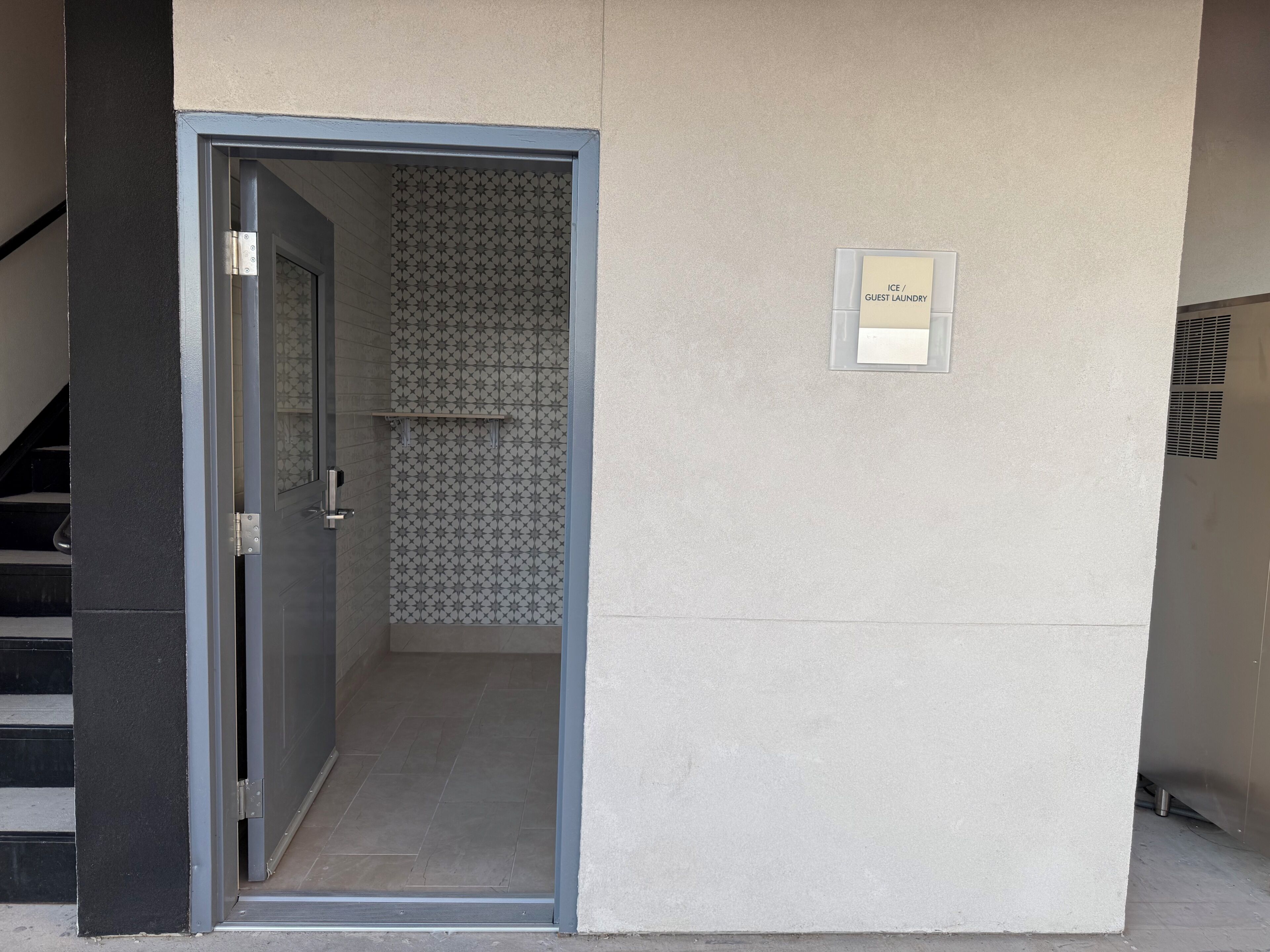 Une entrée d’immeuble avec une porte donnant sur une pièce carrelée, un panneau indiquant la présence d’une buanderie à disposition des clients, et un escalier sur la gauche.