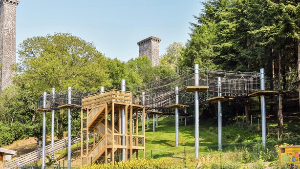 Een houten constructie met een touwbrug, omgeven door groen en een stenen toren.