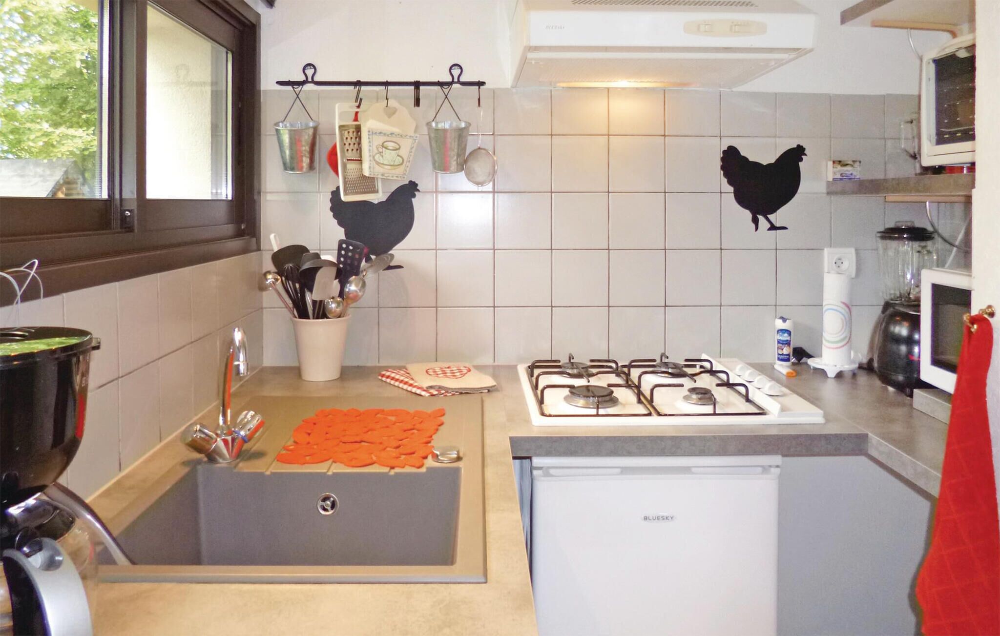 A kitchen with a gas stove, sink, and a refrigerator. There are hanging utensils and a window with a view of trees.
