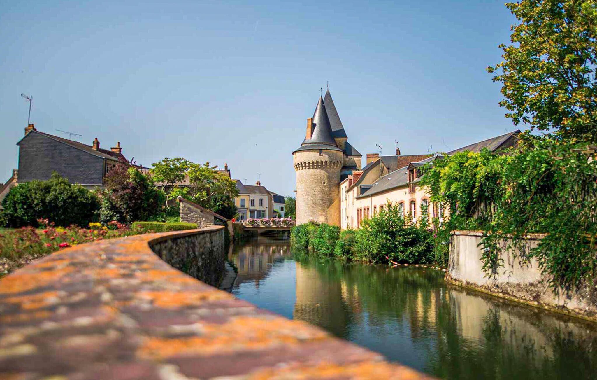 Un canal surmonté d’un pont de pierre, avec une tour et des bâtiments qui bordent la rive.