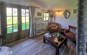 A room with a wooden floor, two sofas with patterned cushions, a coffee table with a teapot, a window with curtains, and a wall-mounted shelf with a vase.