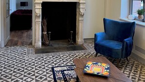 A room with a fireplace, a blue armchair, a patterned rug, and a wooden table with a colorful object on it.