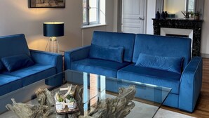 A living room with a blue sofa, a glass coffee table with a tree trunk centerpiece, a chandelier, and a framed painting of a building.