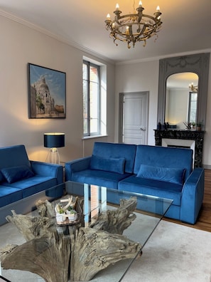 A living room with a blue sofa, a glass coffee table with a tree trunk centerpiece, a chandelier, and a framed painting of a building.