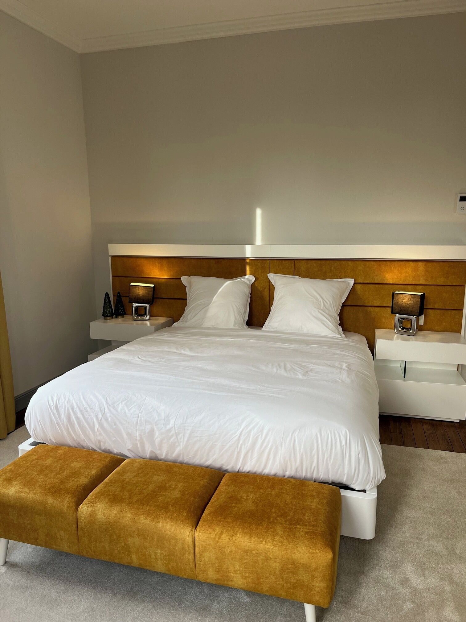 Une chambre à coucher moderne avec un grand lit, des tables de chevet, un banc et une tête de lit dotée d’un éclairage intégré.
