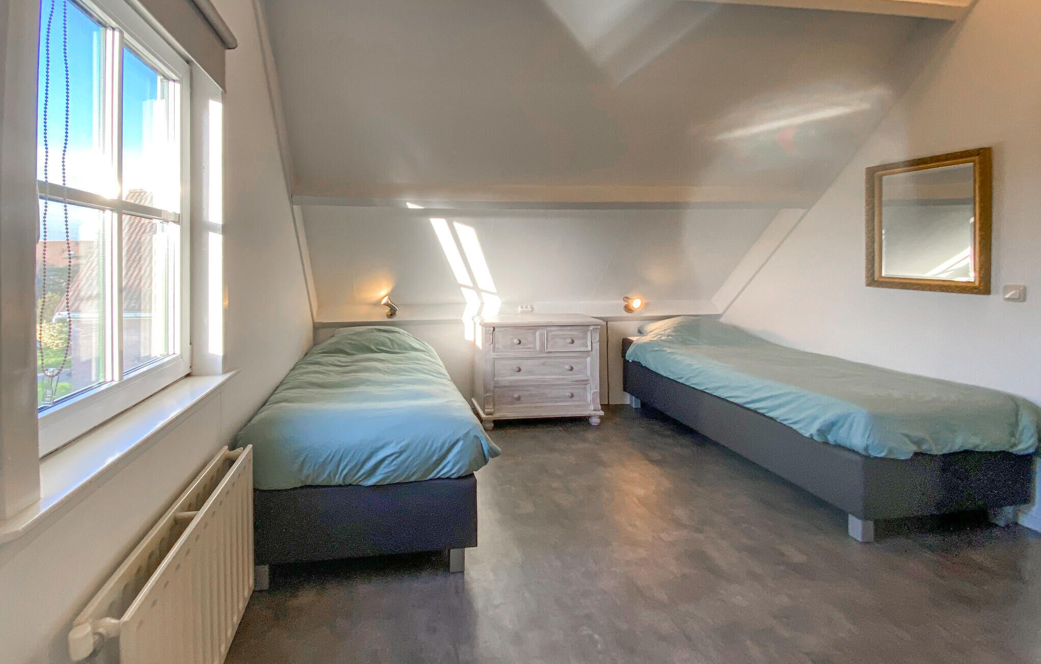 A small, well-lit attic bedroom with two beds, a dresser, and a window with blinds.