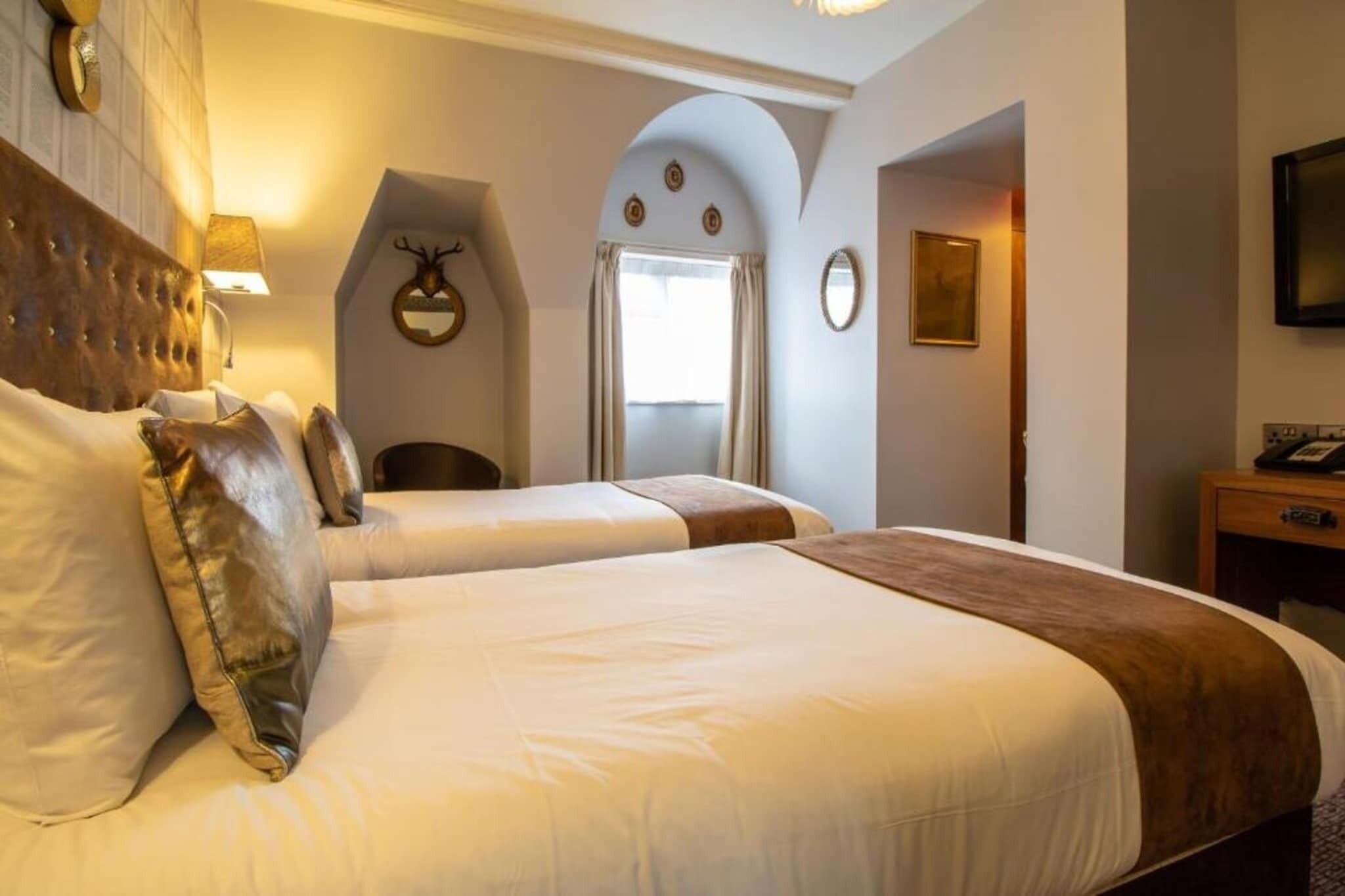A hotel room with two beds, a wooden headboard, a mirror, a wall-mounted TV, and a door leading to another room.