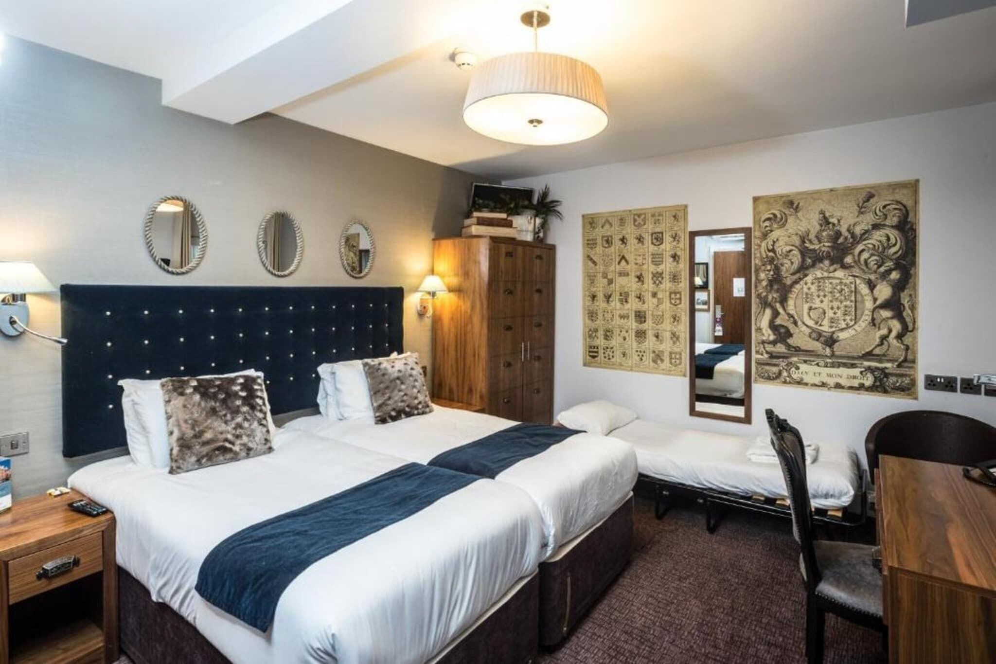A hotel room with two beds, a desk, a chair, a wardrobe, and decorative wall art.