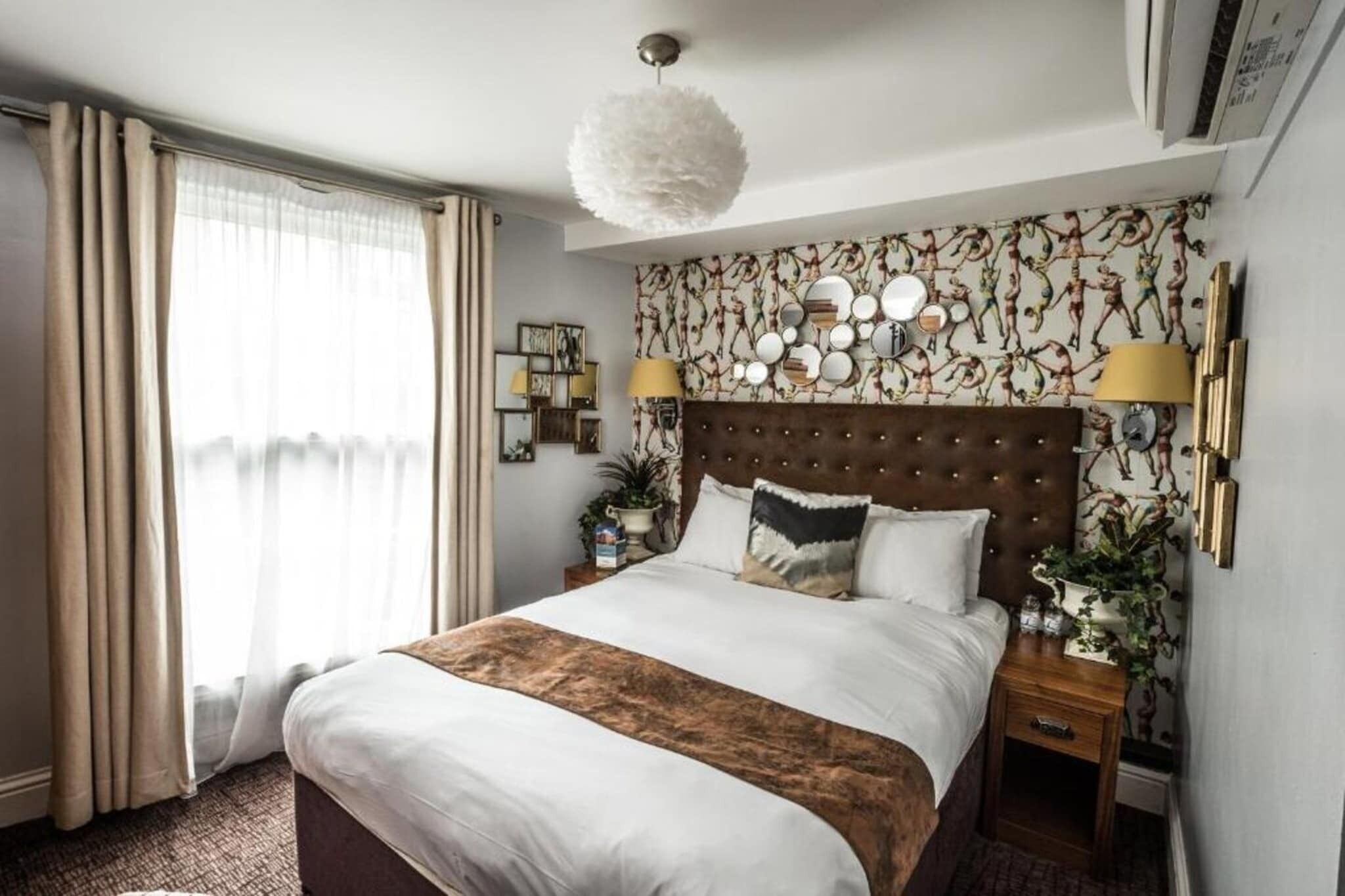 A hotel room with a large bed, a tufted headboard, a chandelier, and a patterned wallpaper.