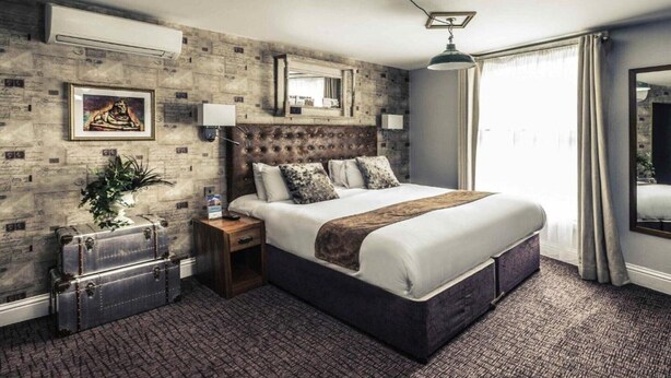 A hotel room with a large bed, a headboard with a mirror, a nightstand, a potted plant, and a suitcase. - Nottingham City Centre Hotel Double Room (Nottingham)
