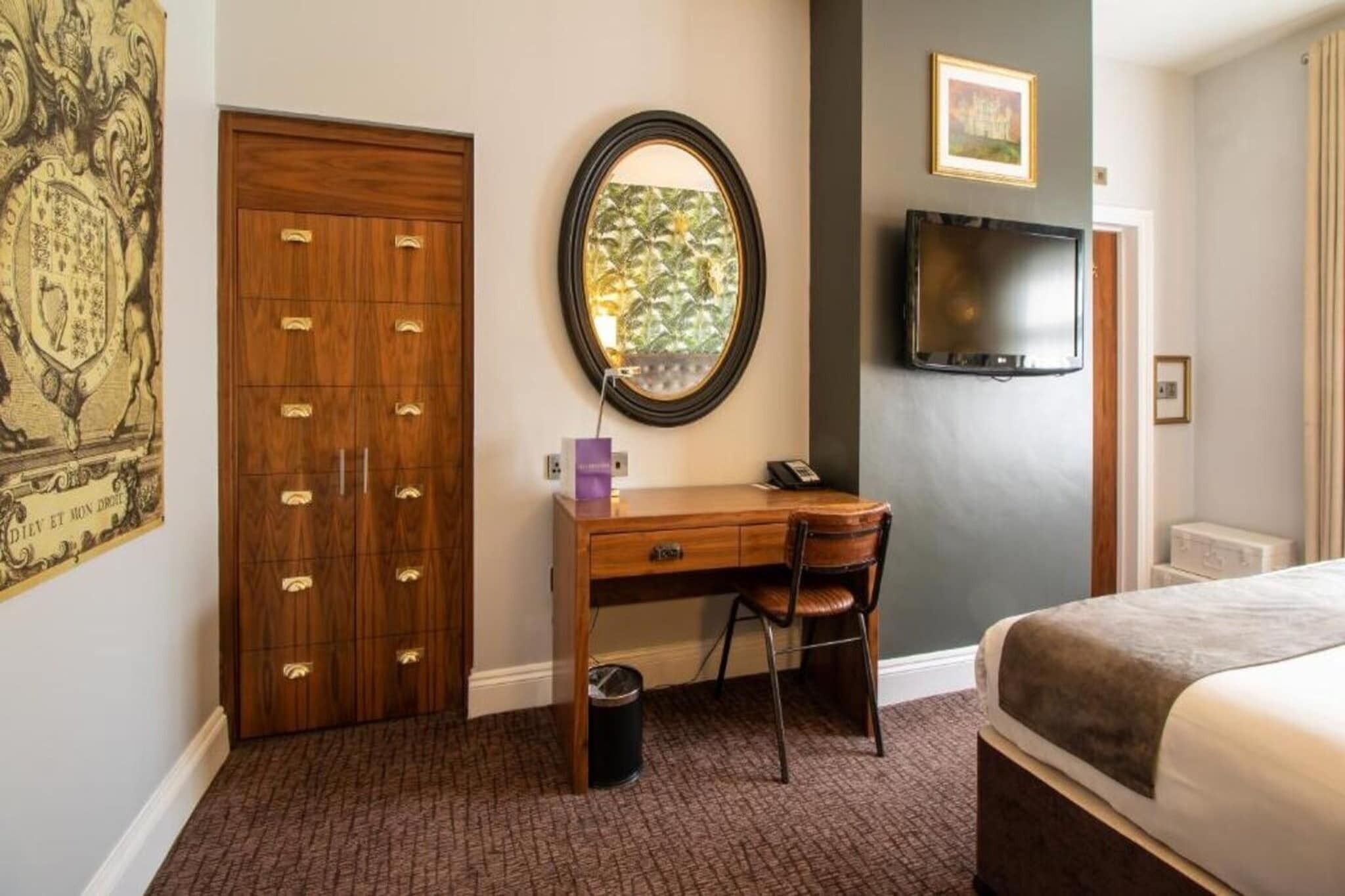 A hotel room with a wooden desk, a chair, a bed, a television, and a wall-mounted mirror.