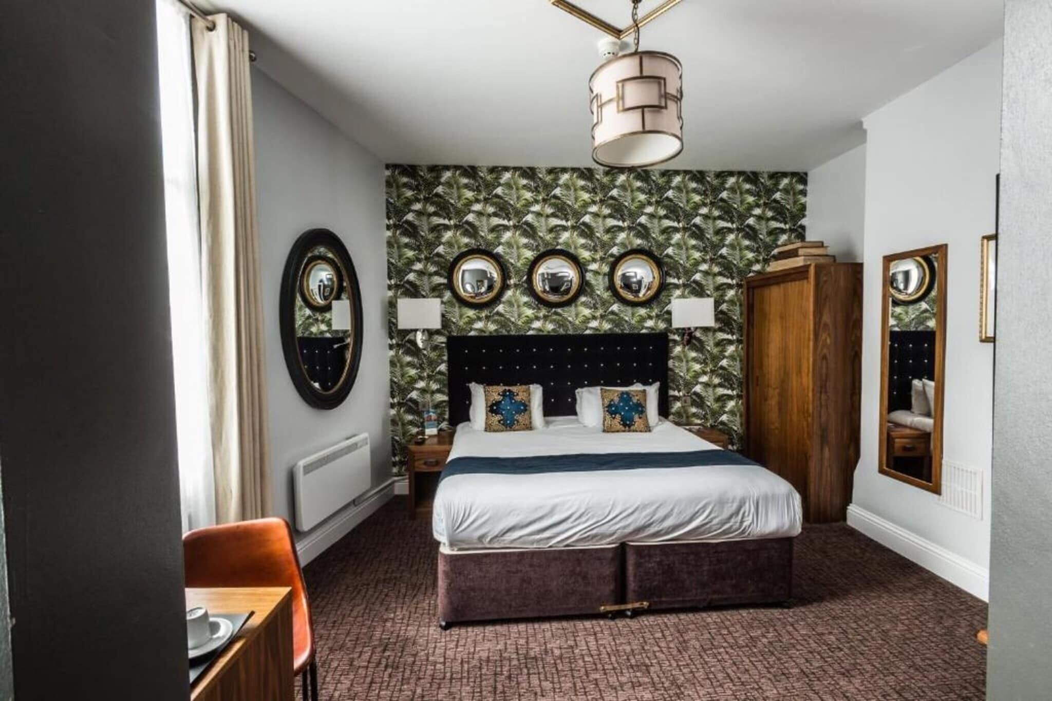 A hotel room with a bed, a wooden wardrobe, a mirror, and a dining area with a table and chairs.