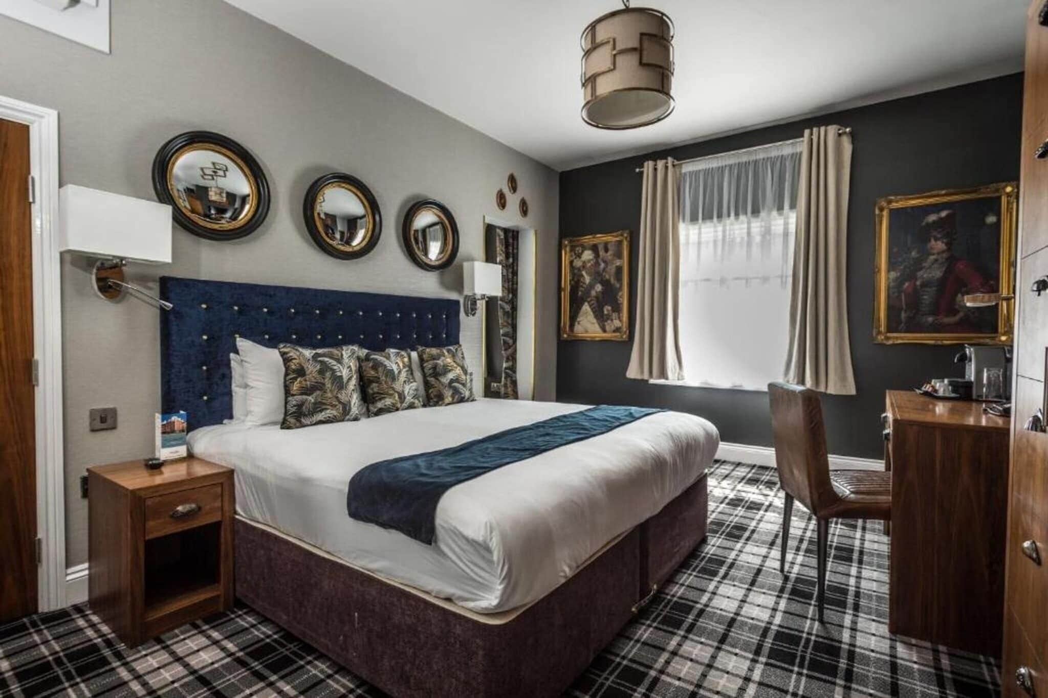 A bedroom with a large bed, a bedside table, a desk, and a chair. There are three wall mirrors, a window with curtains, and a painting on the wall.