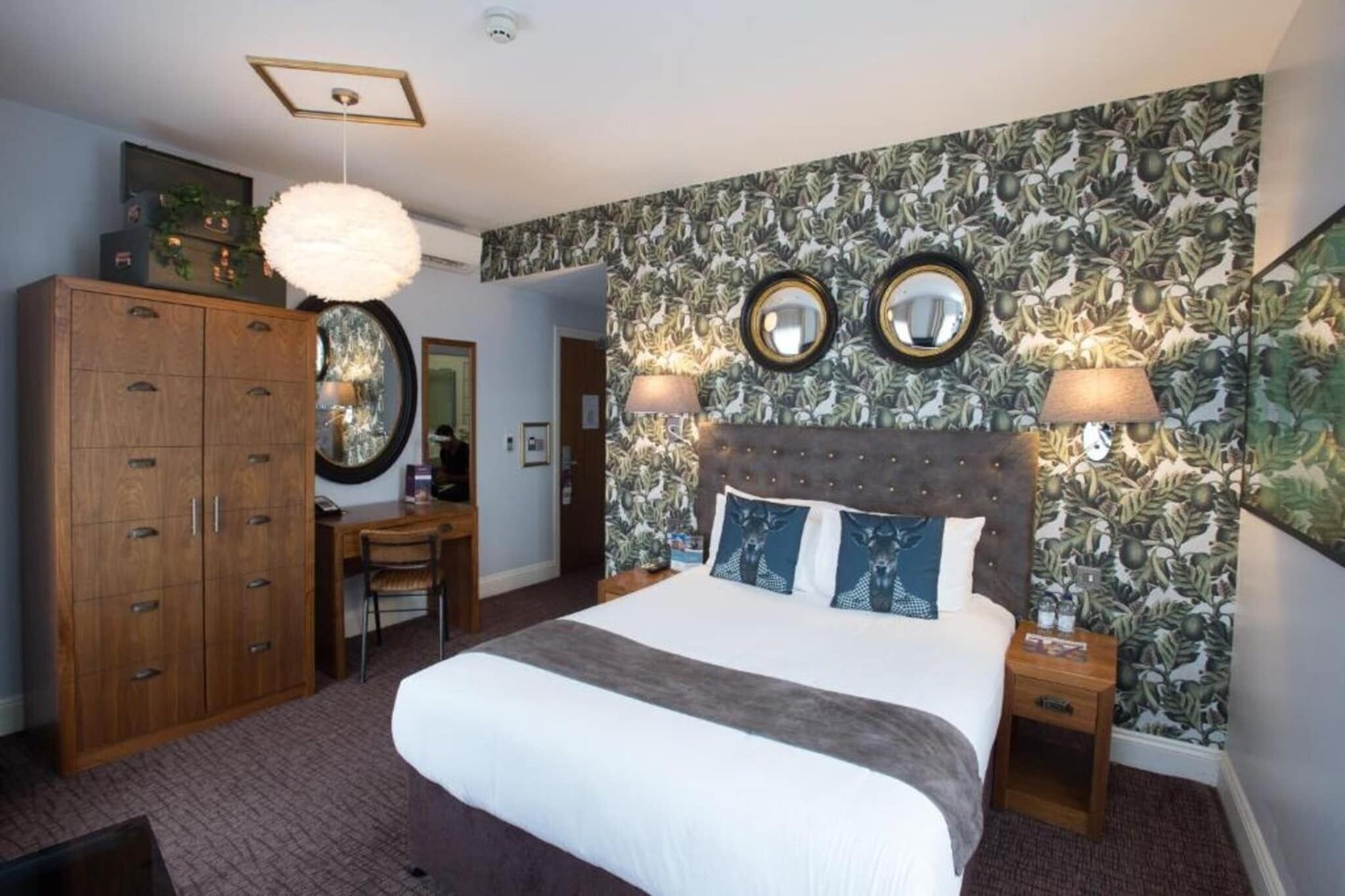 A bedroom with a large bed, two bedside lamps, a wooden wardrobe, a desk, and a mirror on the wall.