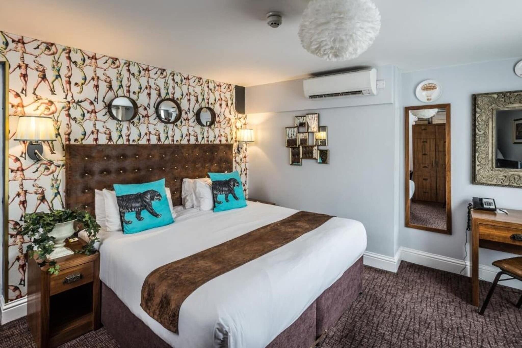 A hotel room with a large bed, a wooden headboard, a nightstand with a lamp, a desk with a chair, and a mirror on the wall.