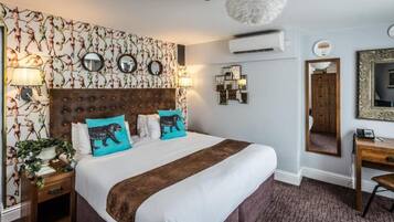 A hotel room with a large bed, a wooden headboard, a nightstand with a lamp, a desk with a chair, and a mirror on the wall.