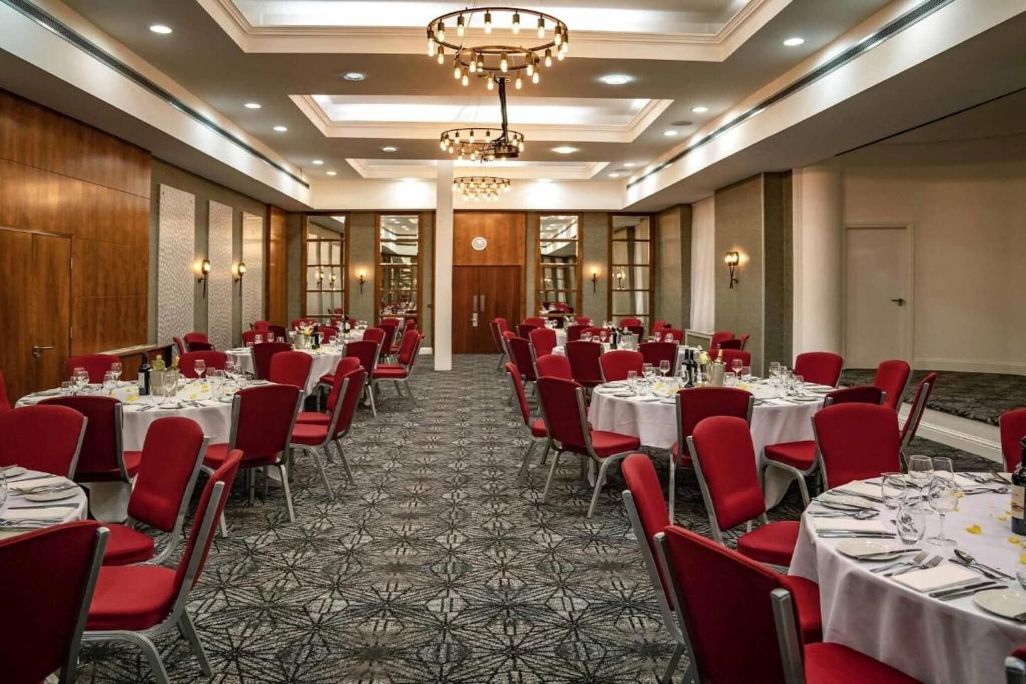 A banquet hall with round tables, red chairs, and a patterned carpet.