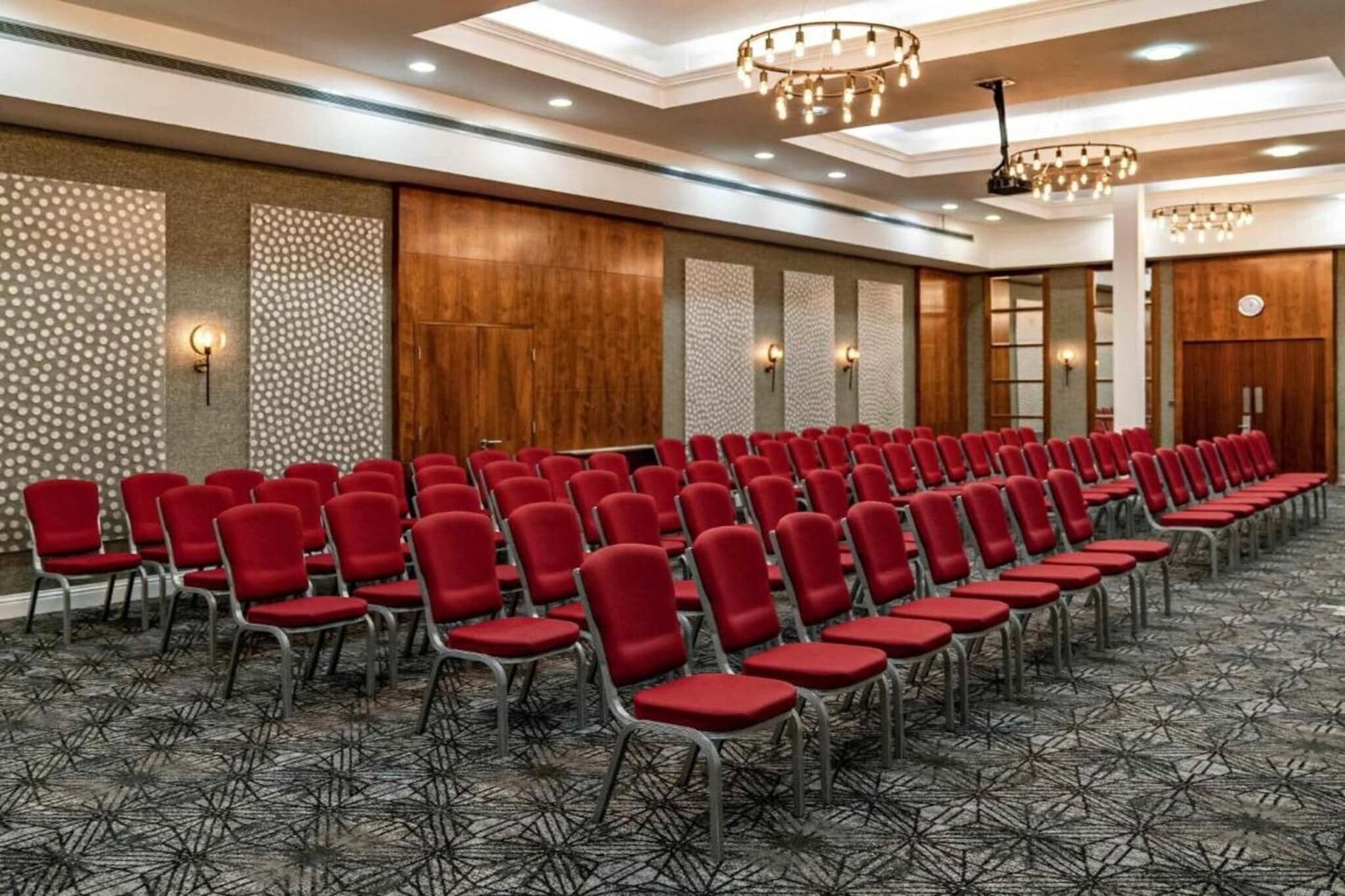 A conference room with rows of red chairs, patterned carpet, wooden paneling, and wall sconces.
