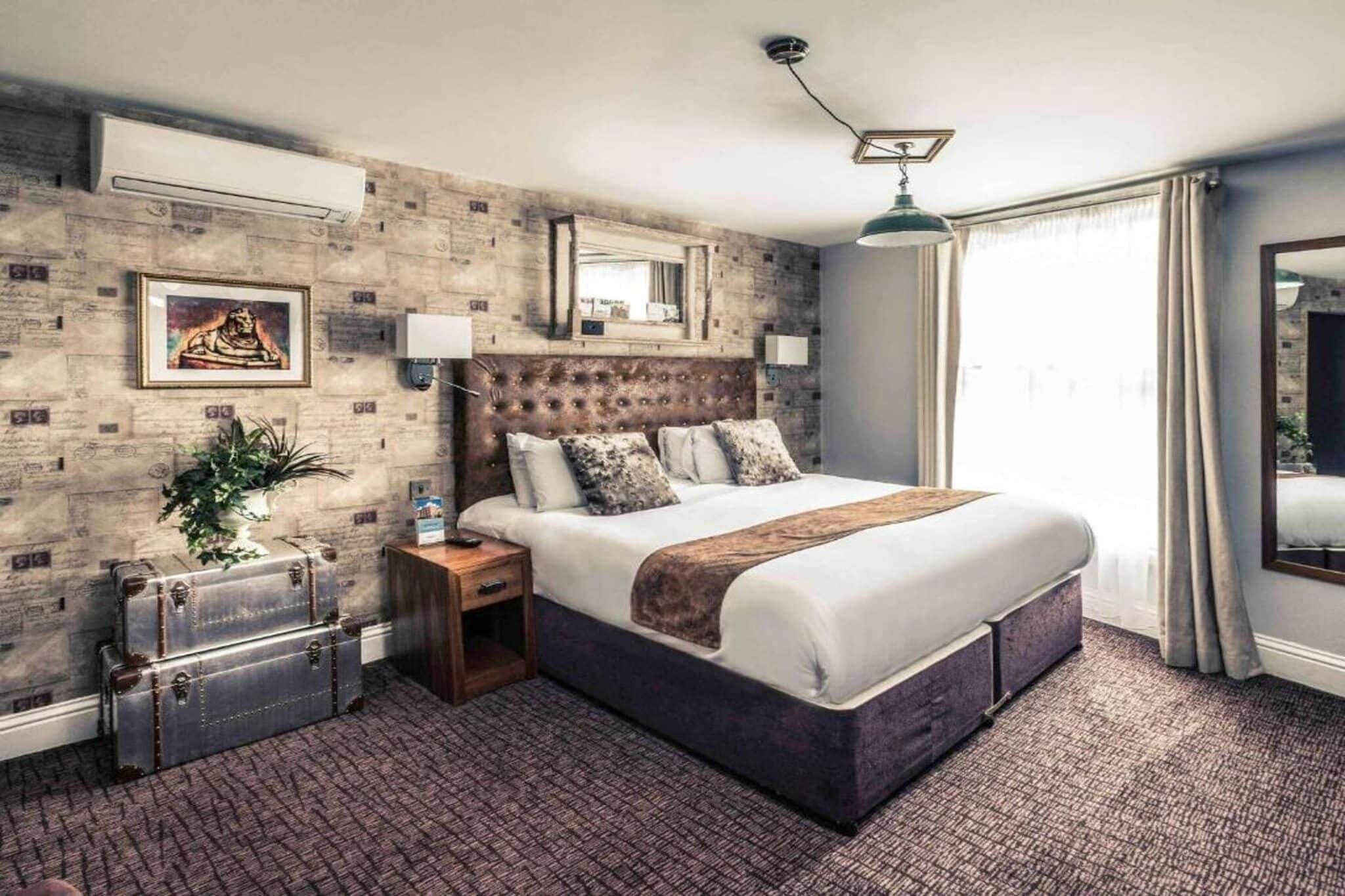 Een hotelkamer met een groot bed, een nachtkastje, een spiegel, een bloempot en een koffer.