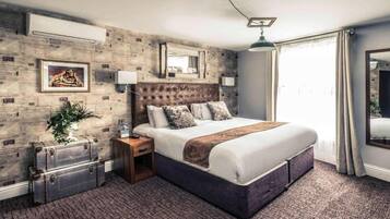 A hotel room with a large bed, a nightstand, a mirror, a potted plant, and a suitcase.