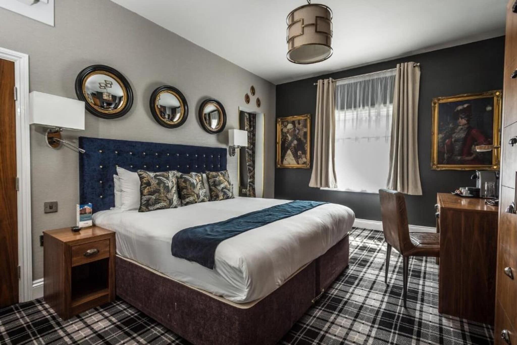 Une chambre avec un grand lit, une table de chevet, un bureau et une chaise. On y trouve trois miroirs muraux, une fenêtre avec des rideaux et un tableau accroché au mur.
