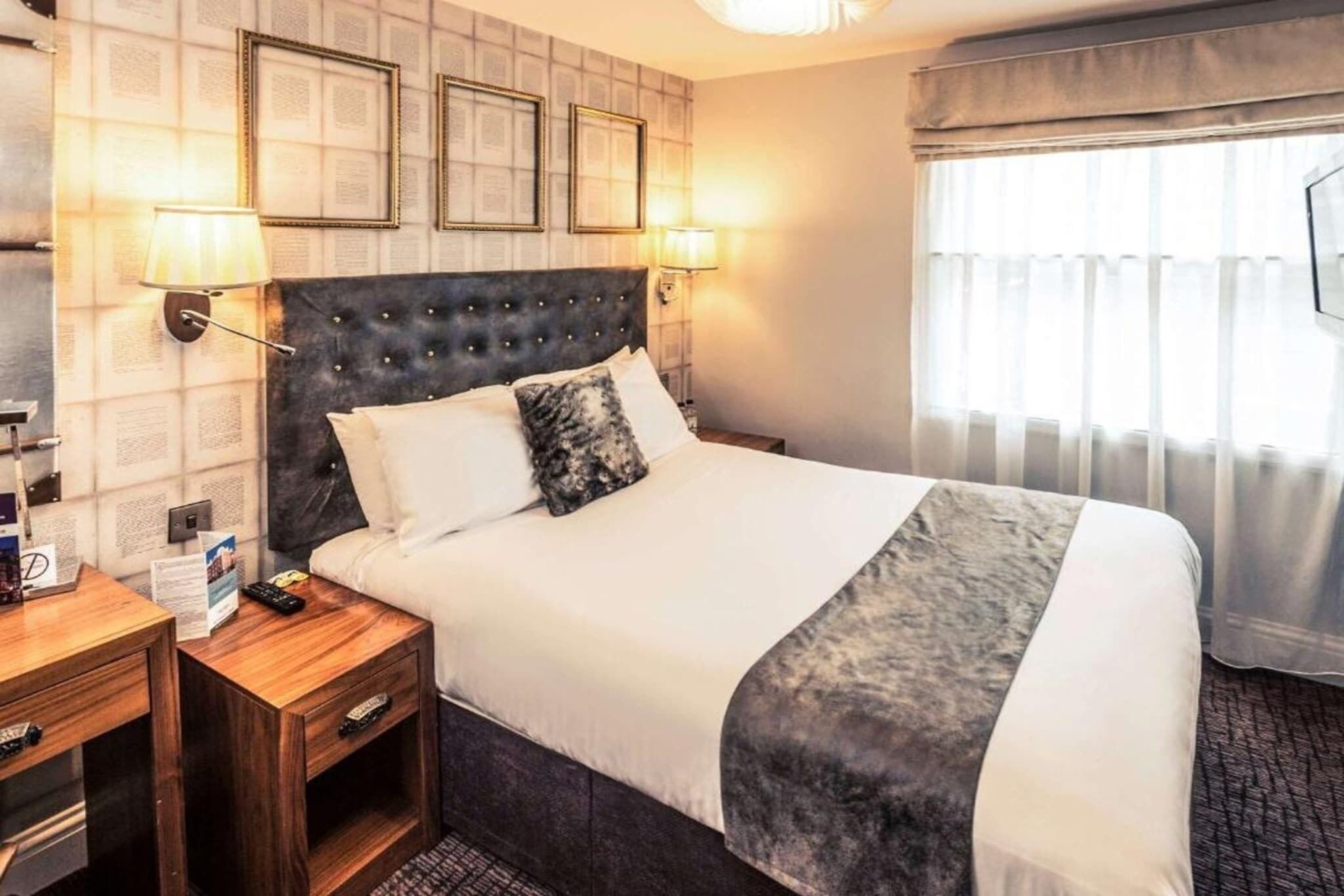A hotel room with a large bed, two bedside tables, a nightstand, a wall-mounted lamp, and a window with curtains.