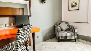 Een kamer met een fauteuil met een hondentand patroon, een televisie op een oranje tafel, een wandlamp en een ingelijst kunstwerk aan de muur.