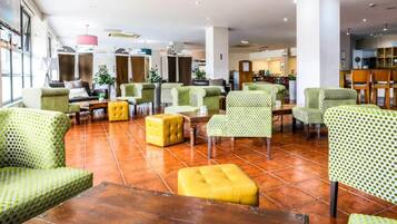Un hall d’entrée spacieux doté de plusieurs espaces de détente, notamment des fauteuils à motifs verts et des poufs jaunes, et une cuisine ouverte visible au fond.