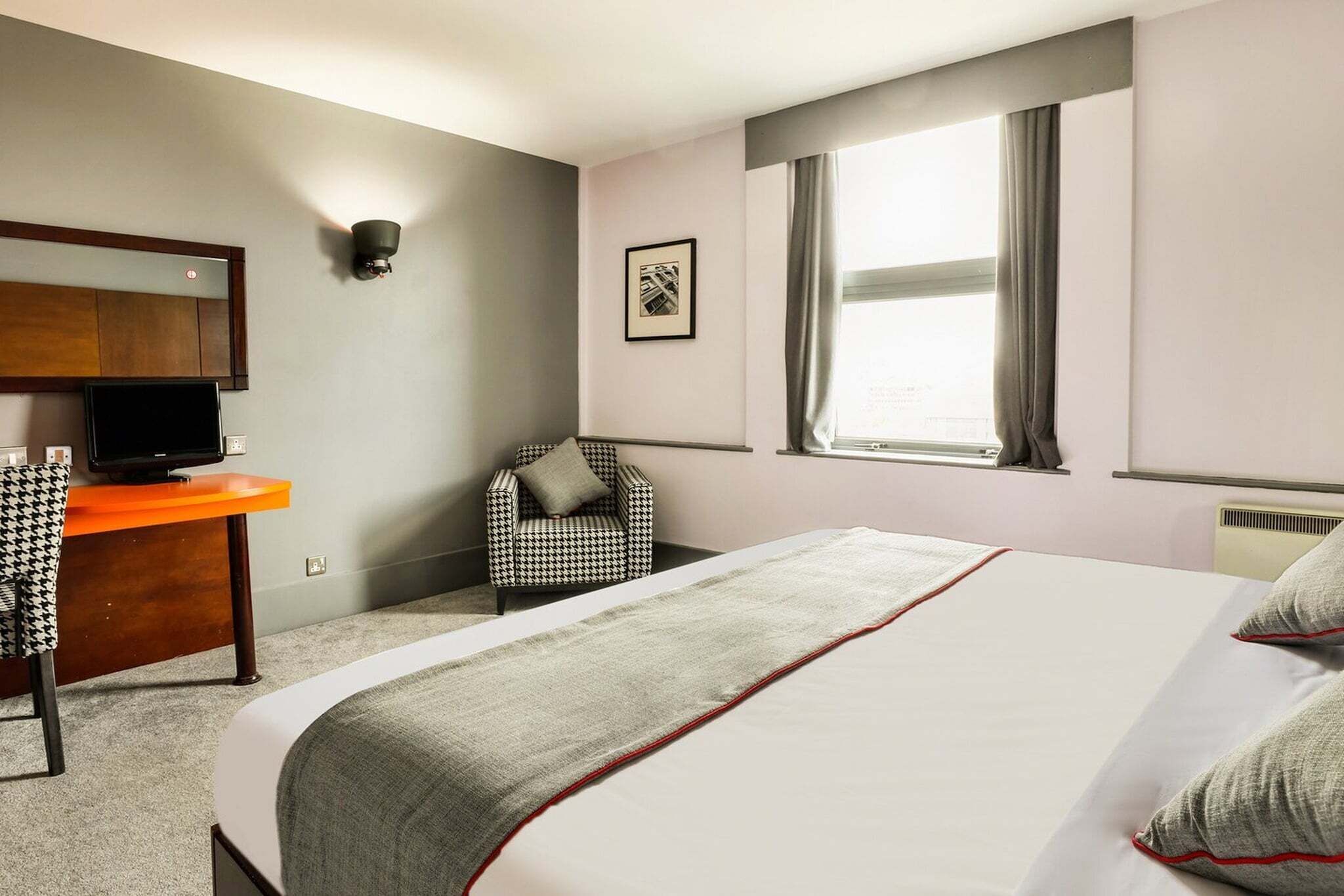 Une chambre d’hôtel comprenant un lit, un bureau avec une télévision, une chaise, une fenêtre avec des rideaux et un luminaire fixé au mur.