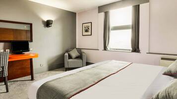 Una habitación de hotel con cama, escritorio con televisor, silla, ventana con cortinas y una lámpara fijada a la pared.