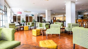 Un vestíbulo amplio con varias zonas de estar, incluyendo sillones estampados verdes y otomanas amarillas, y una cocina visible al fondo.