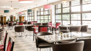 Un interior de restaurante con varias mesas y sillas, ventanas amplias y lámparas colgantes.