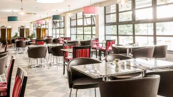 L’intérieur d’un restaurant, avec de nombreuses tables et chaises, de grandes fenêtres et des luminaires suspendus.
