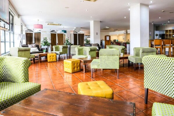 A spacious lobby with multiple seating areas, including green patterned chairs and yellow ottomans, and a visible kitchen area in the background.