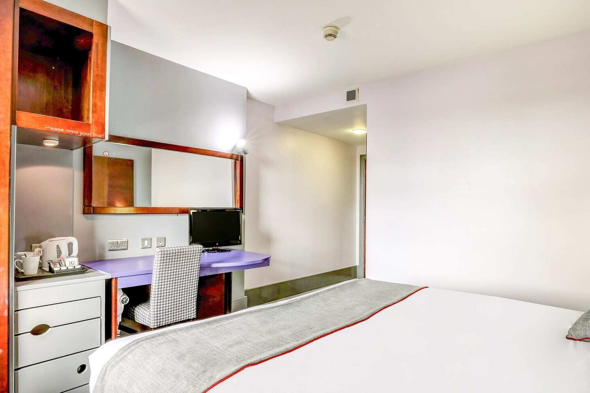 Et hotelværelse med seng, skrivebord med computer, stol, lille bord med elkedel og et spejl.