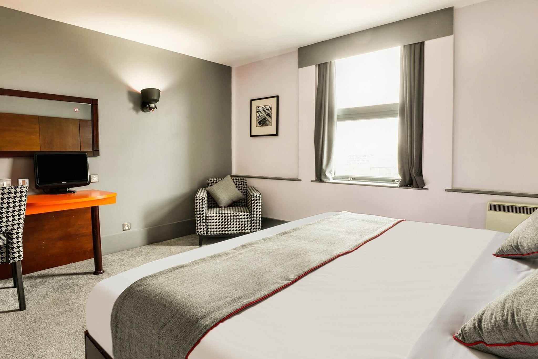 A hotel room with a bed, a desk with a television, a chair, a window with curtains, and a wall-mounted light fixture.
