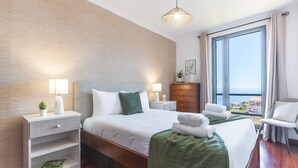A hotel room with a bed, bedside tables, a dresser, a chair, and a large window with a view. - Aurora Atlantic Apartment by Atlantic Holiday (Caniço)