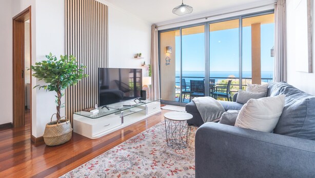 A modern living room with a flat-screen TV, a grey sofa, a small round table, and a large window offering a view of the sea. - Aurora Atlantic Apartment by Atlantic Holiday (Caniço)