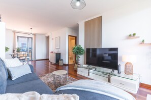 Ein modernes Wohnzimmer mit Sofa, Fernseher, Glastisch und einem Essbereich mit Stühlen und Tisch.