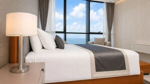 A modern hotel room with a large bed, a bedside lamp, a view of the sky through large windows, and a view of the sea.