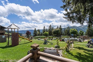A playground with a seesaw, climbing wall, and slide, surrounded by trees and a lake.