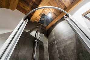 A shower with a glass door, a wooden ceiling, and dark tiled walls.