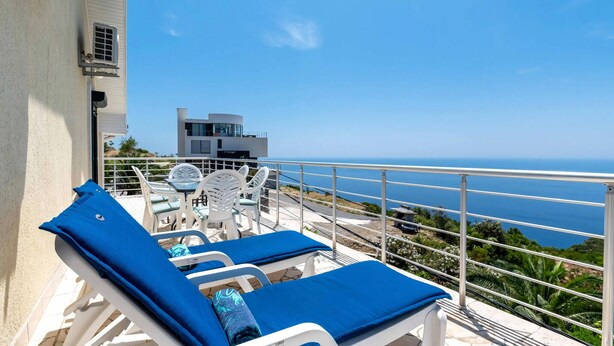 Un balcon doté de chaises longues et d’une table, offrant une vue sur la côte, avec un bâtiment moderne en arrière-plan.