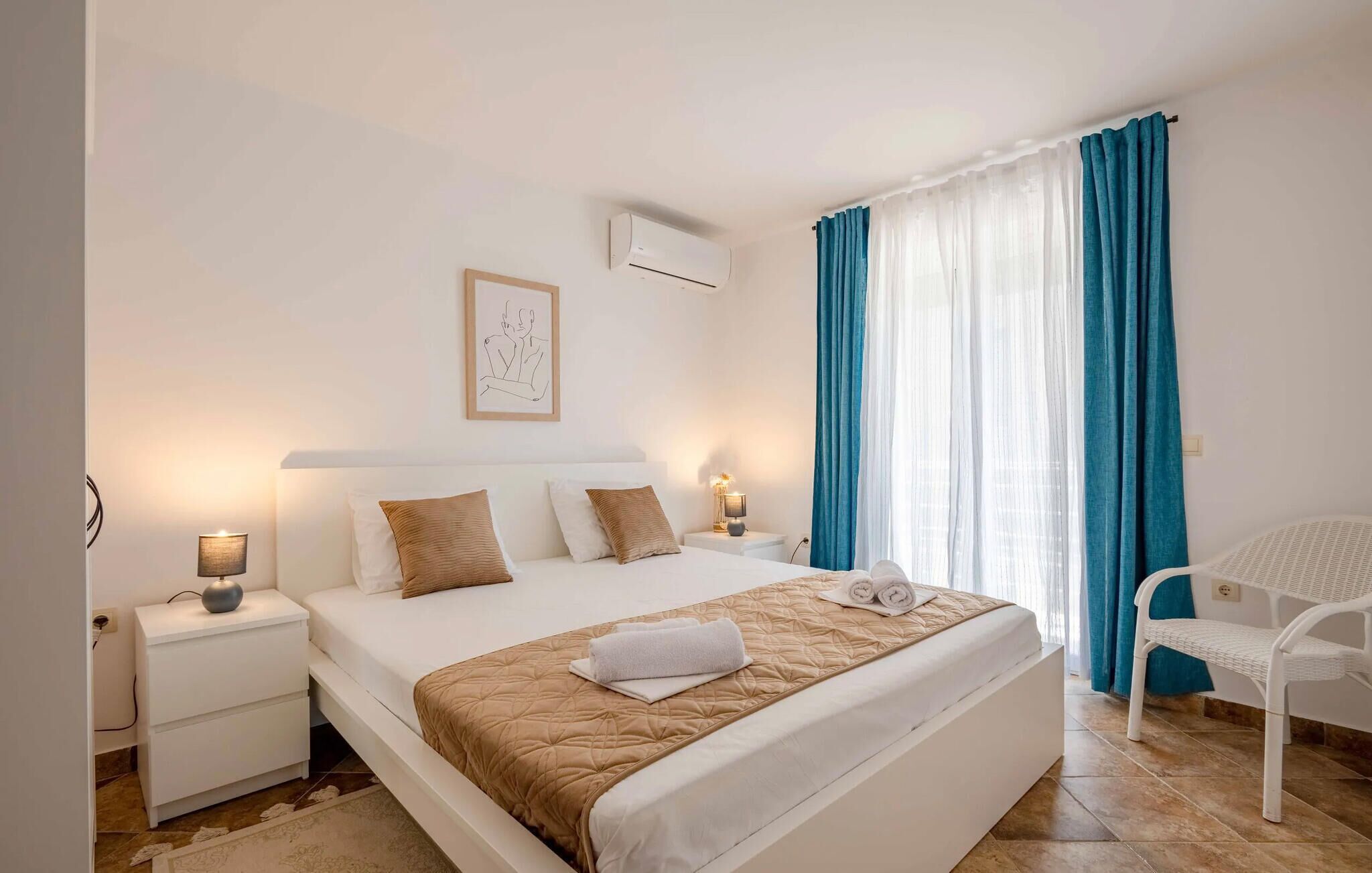 Una habitación de hotel con cama, mesitas de noche, aire acondicionado en la pared, una ventana con cortinas azules y una silla blanca.