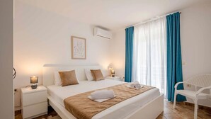 Et hotelværelse med en seng, sengeborde, en fastgjort aircondition, et vindue med blå gardiner og en hvid stol.